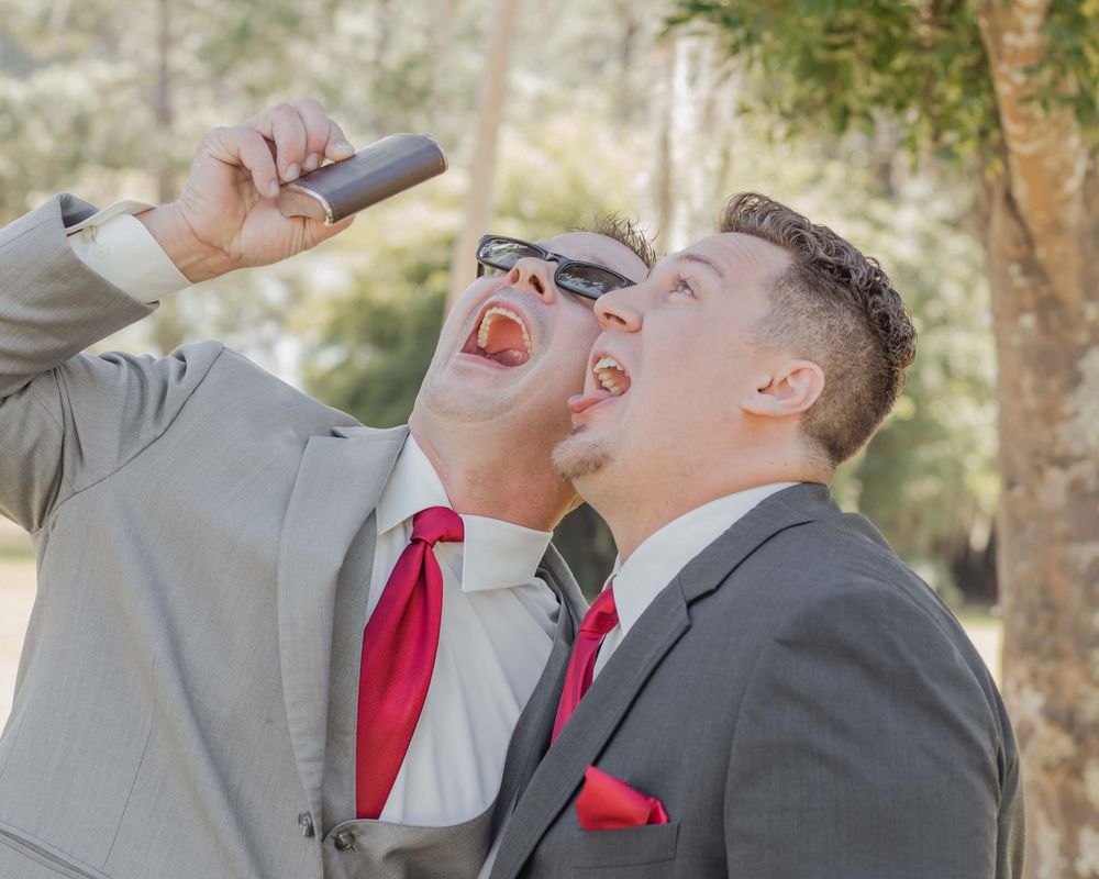 Two groomsmen in gray suits with red ties playfully pose while drinking from flasks at an outdoor wedding celebration.