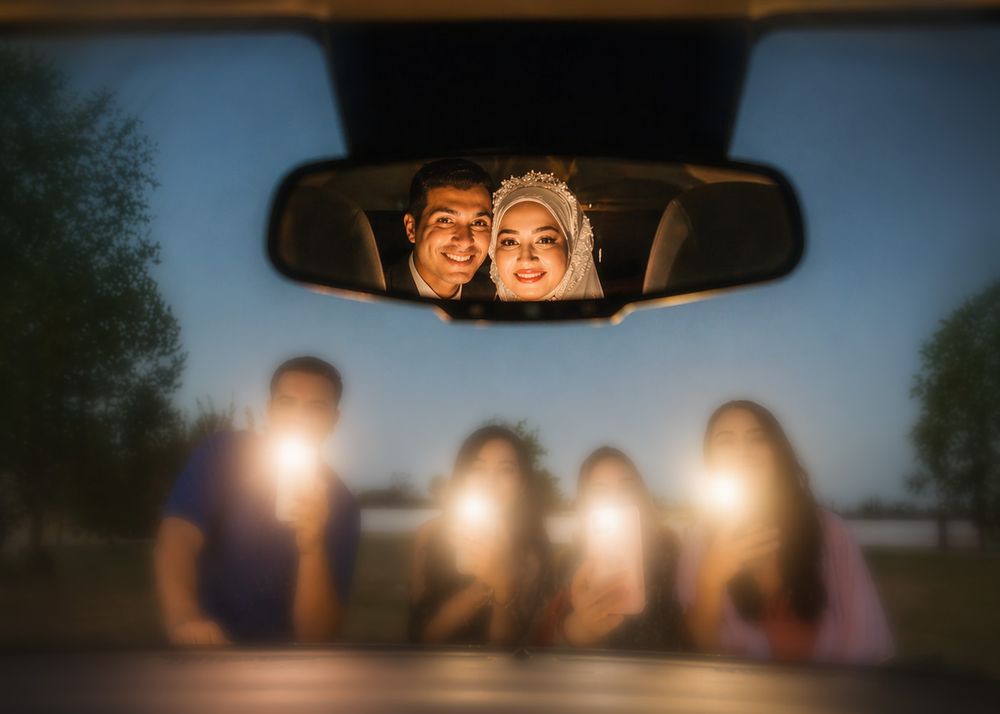 Couple reflected in car rearview mirror during wedding night, with blurred guests holding lights in background.