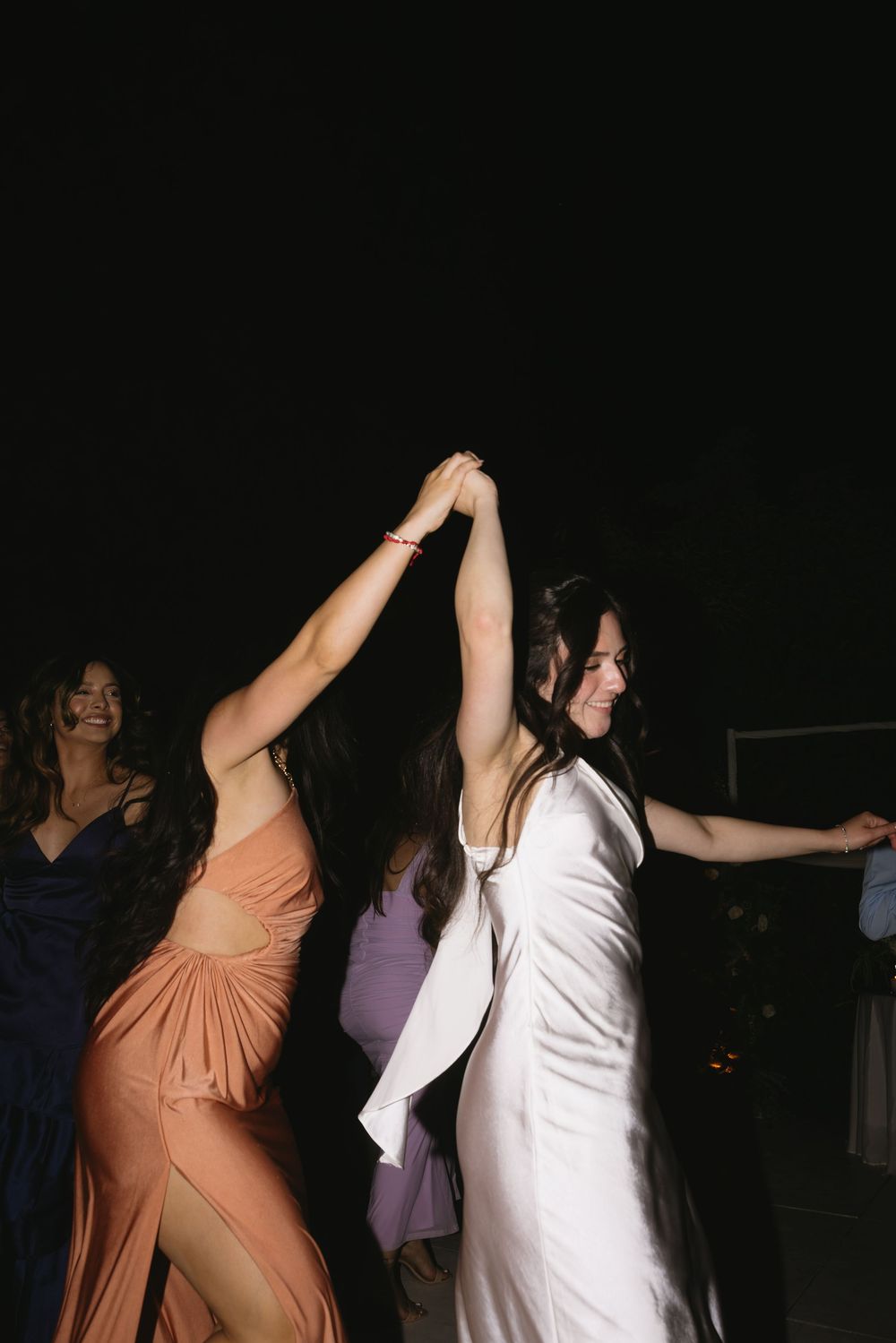 Two women dancing together at a party wearing elegant dresses in peach and white colors against a dark background.