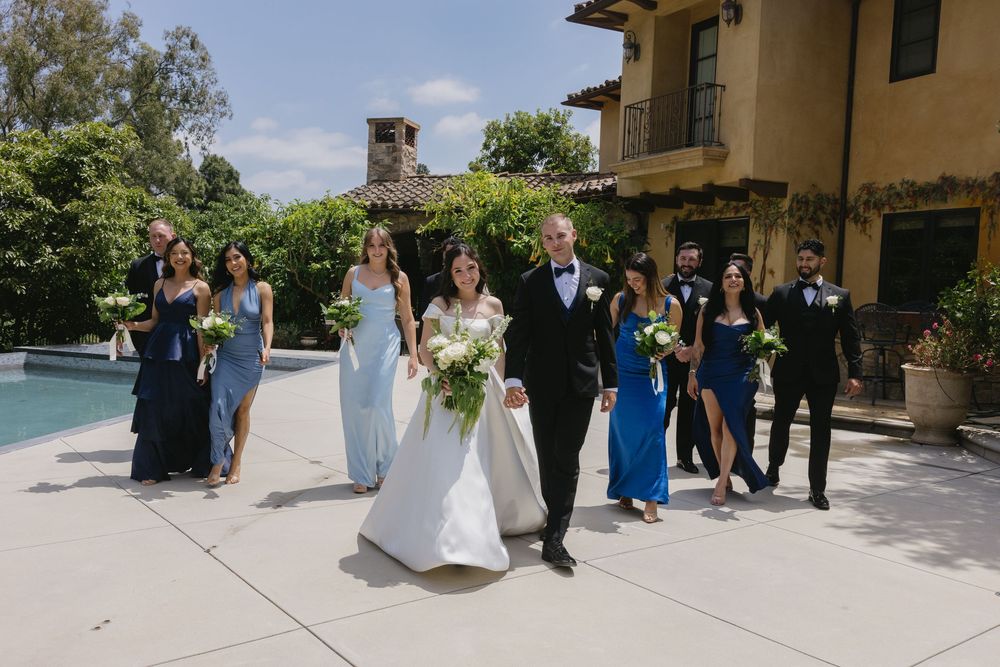 Wedding party gathers outdoors on stone patio surrounded by lush landscaping during sunny daytime celebration.