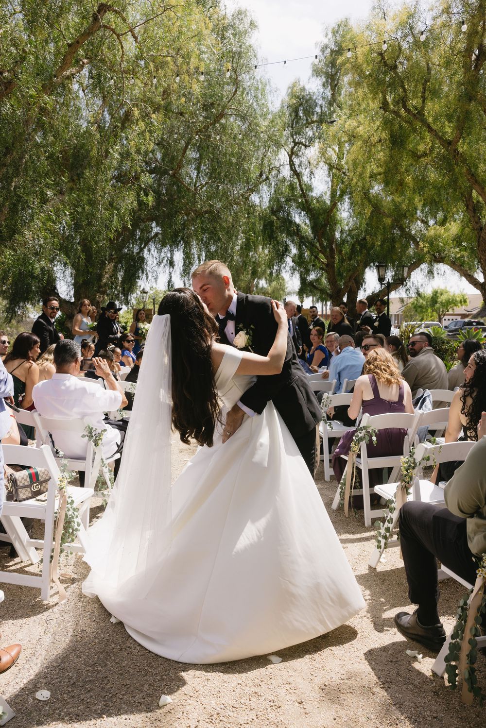 A couple dances during an outdoor wedding ceremony under willow trees while guests watch from white folding chairs.