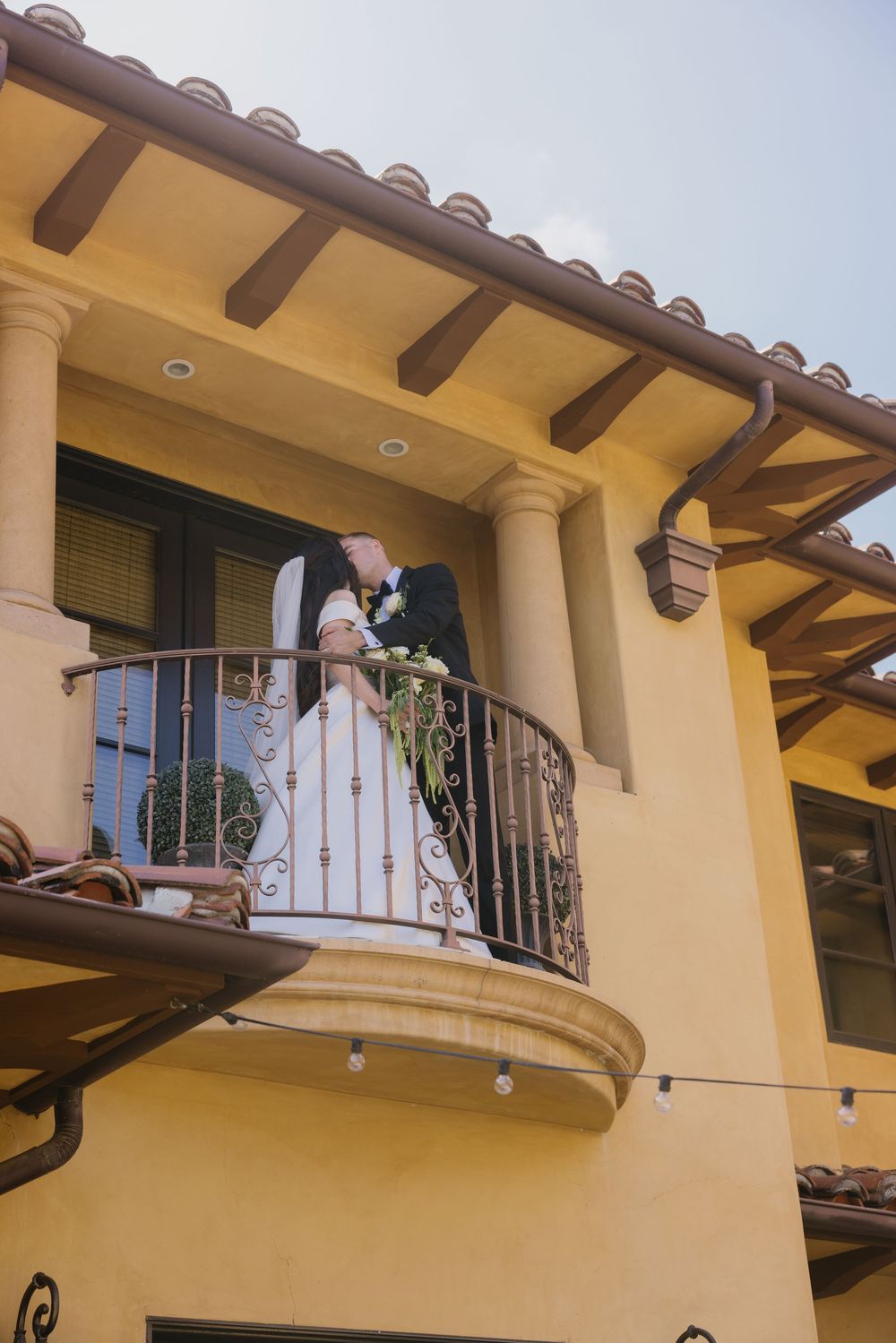 Two people share a romantic moment on a Mediterranean-style balcony with yellow stucco walls and Spanish tile roof details.
