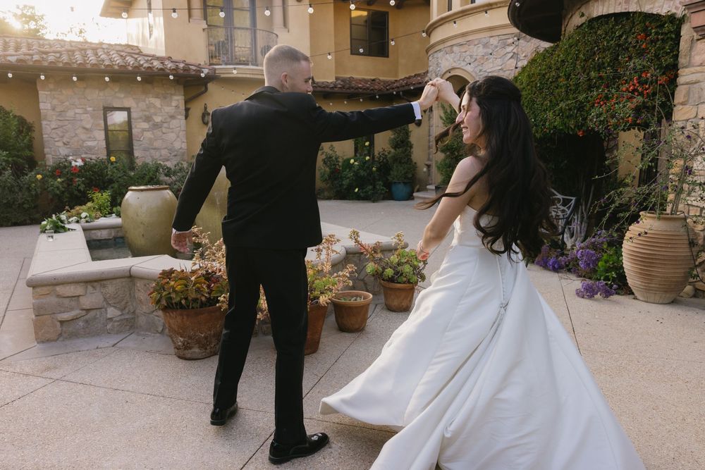 A newlywed couple dancing together on an outdoor stone patio surrounded by potted plants and garden decor.