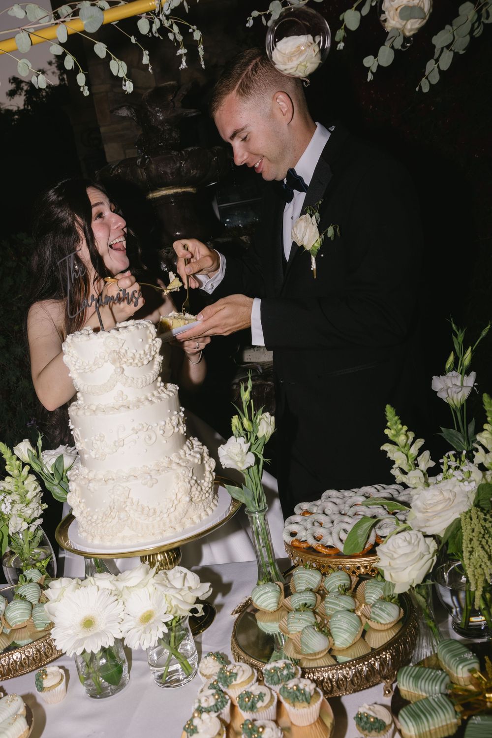 A couple share a playful moment cutting their elegant white wedding cake surrounded by cupcakes and floral decorations.