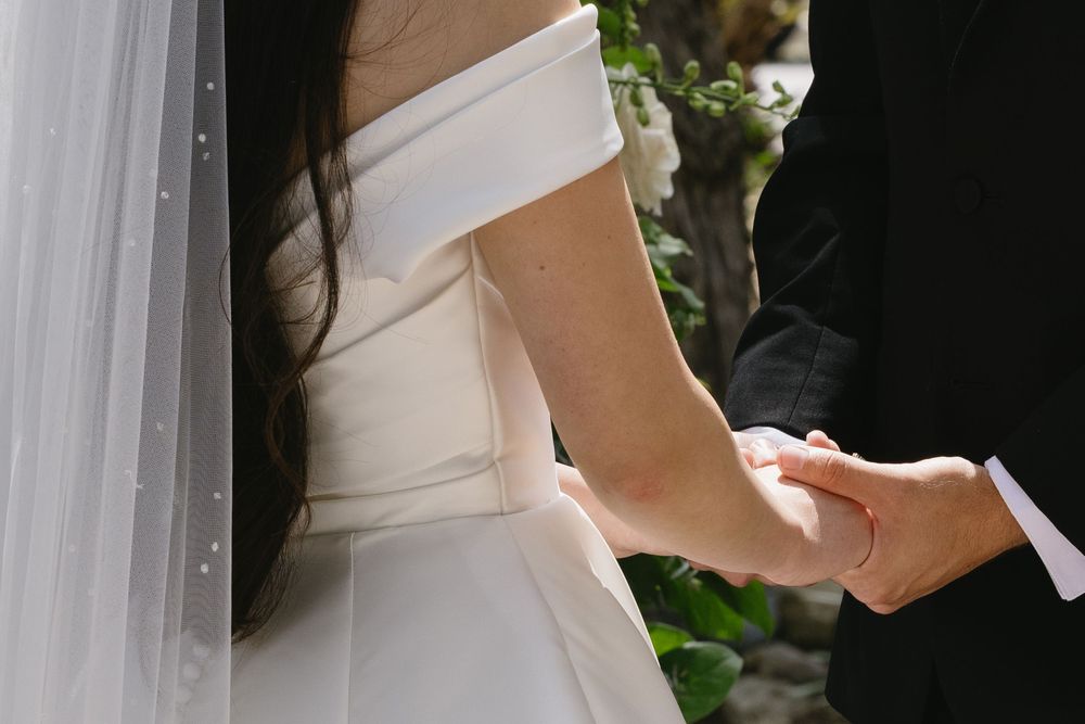 Hands clasped together during a wedding ceremony, with a white off-the-shoulder bridal gown and veil visible.