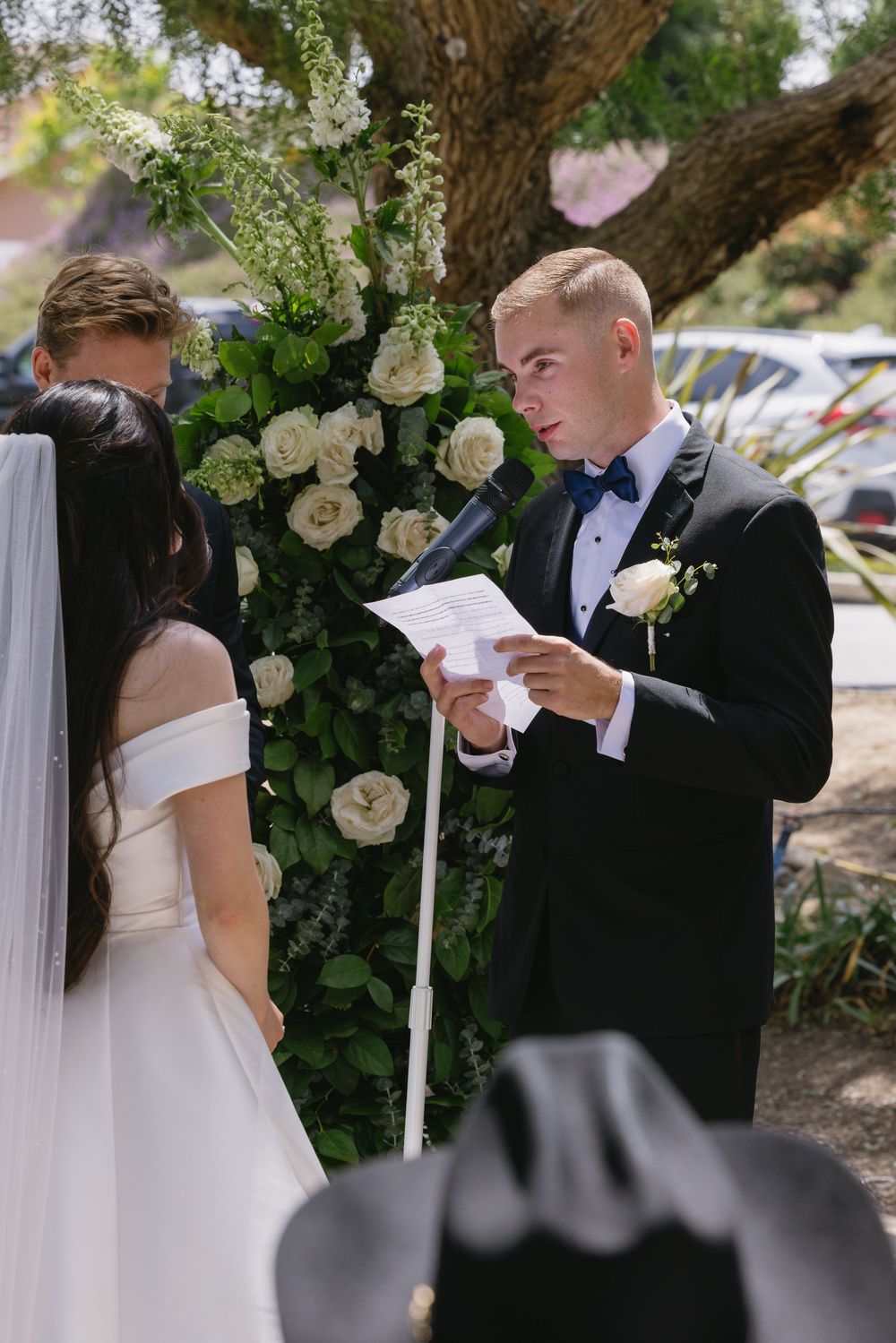 An outdoor wedding ceremony under trees with white roses and greenery decorating the altar area.