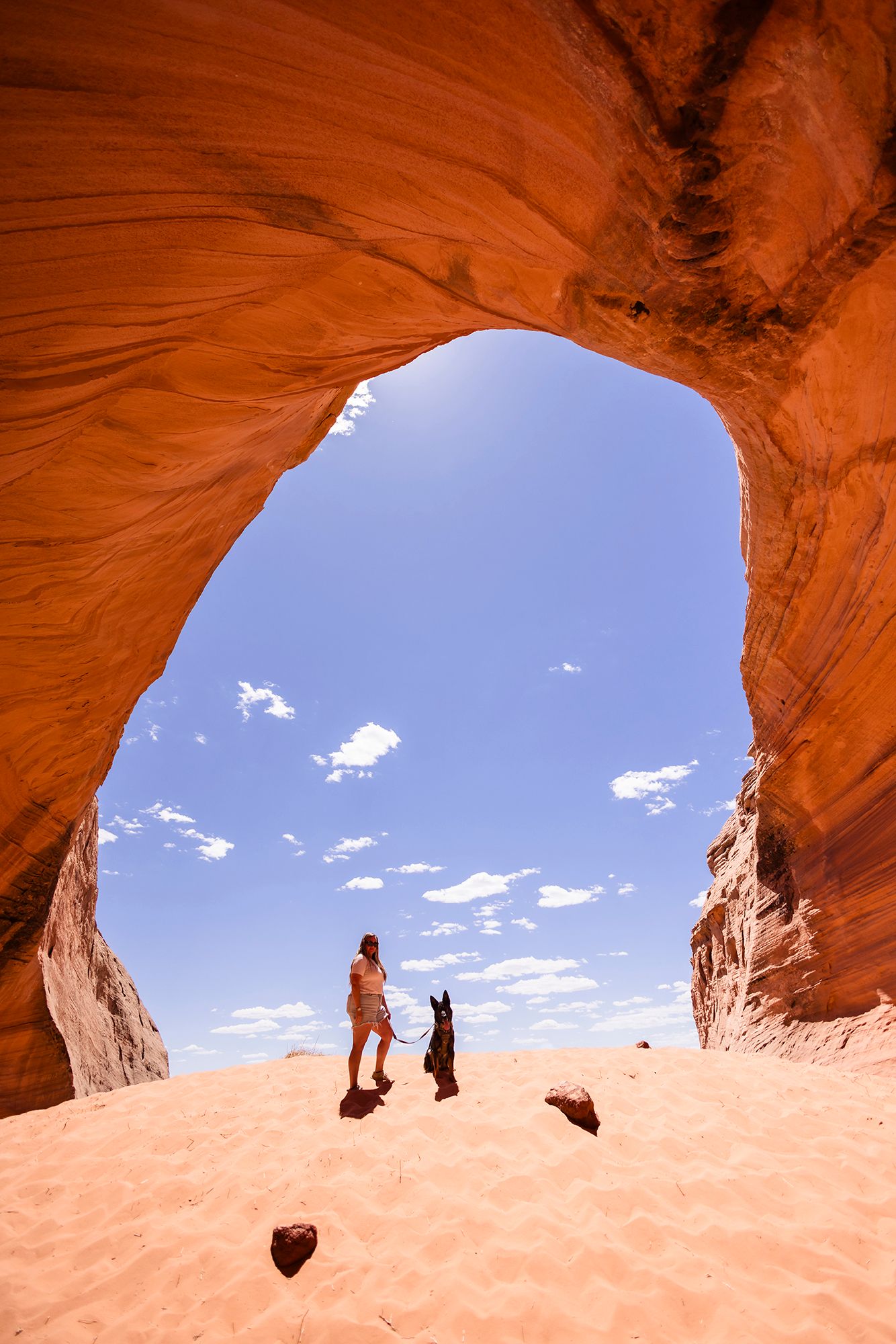 Two silhouettes stand beneath a natural rock arch formation at Arches National Park against a bright blue sky with clouds.
