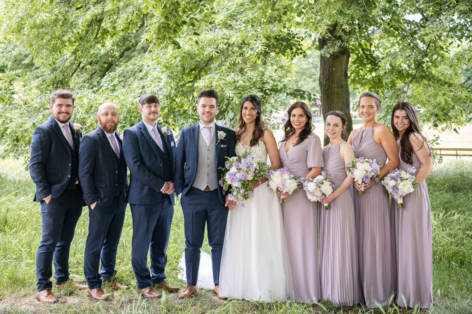 Group wedding photo with four groomsmen next to groom and four bridesmaids in lilac dresses next to bride. Standing under tree in park.