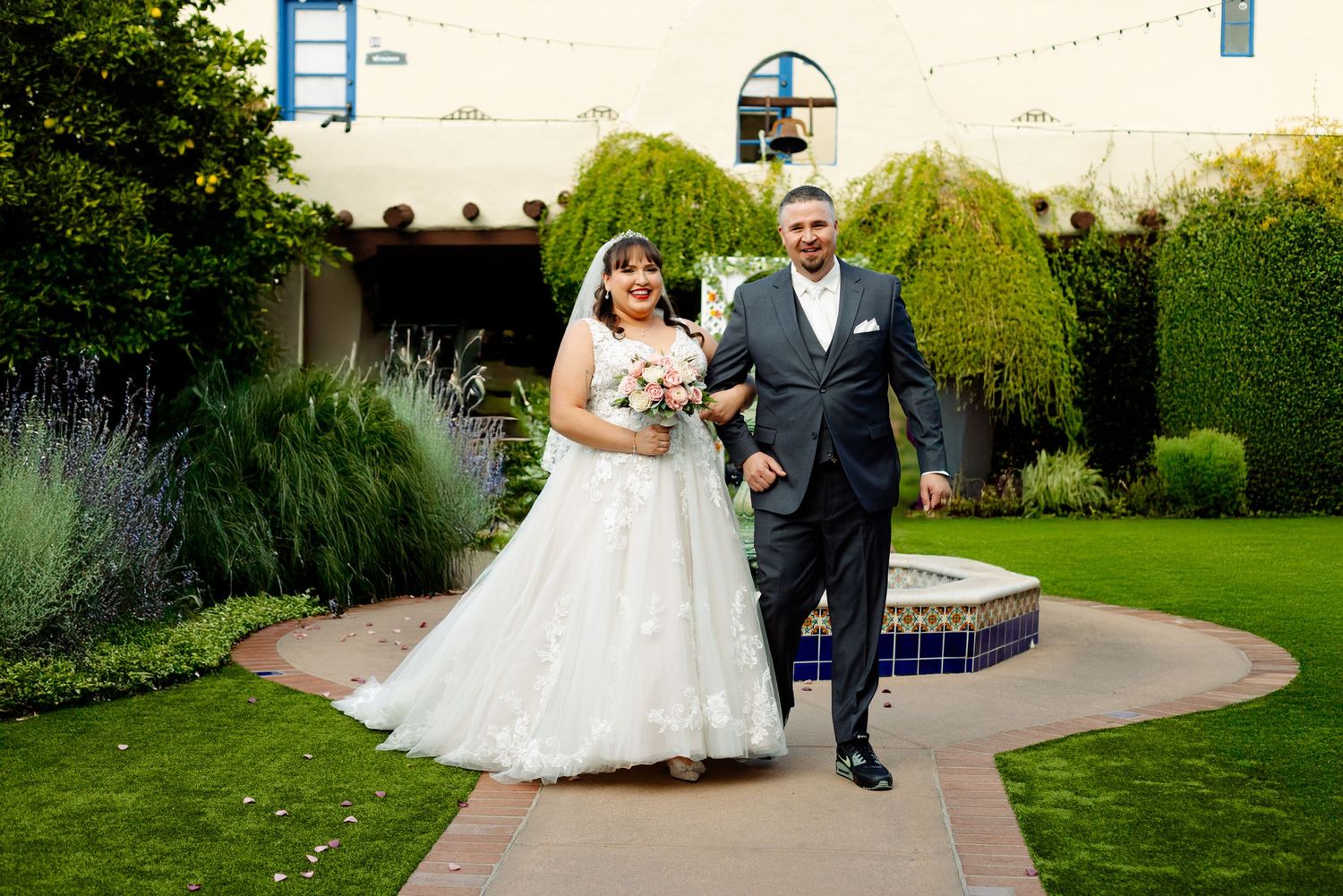 A couple shares an intimate moment along a tree-lined garden pathway with soft natural lighting at dusk.