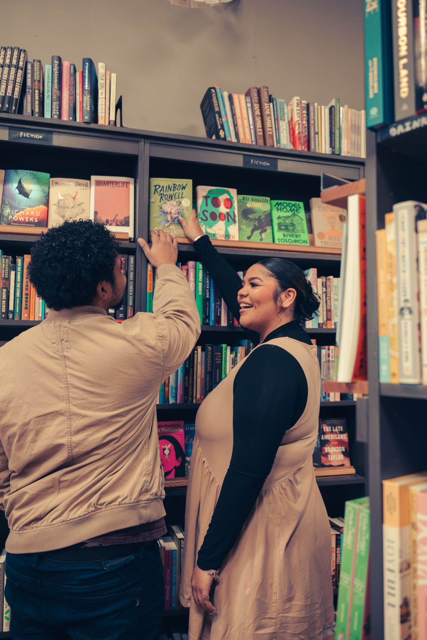 Two people browsing books together at a bookstore, reaching for titles on wooden shelves while sharing a joyful moment.