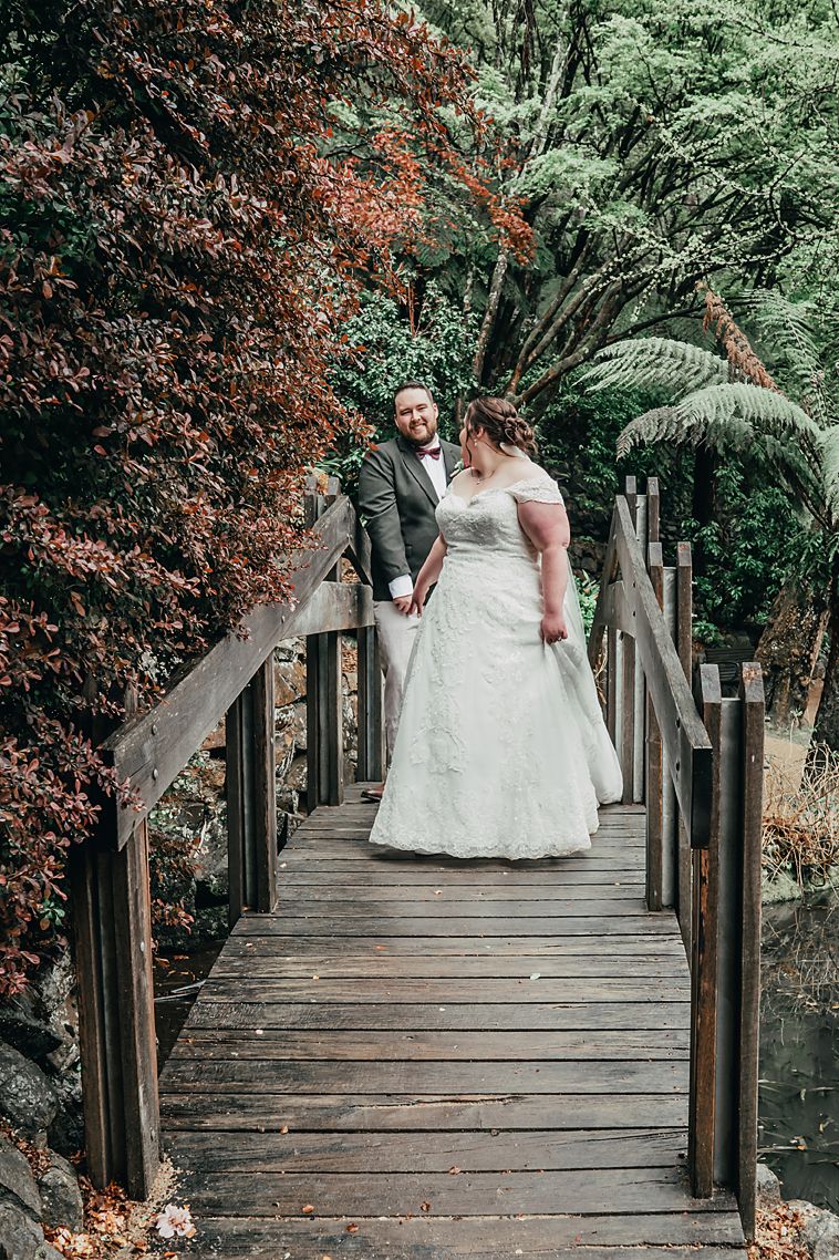A couple stands together on a rustic wooden bridge surrounded by lush greenery and red foliage in a garden setting.