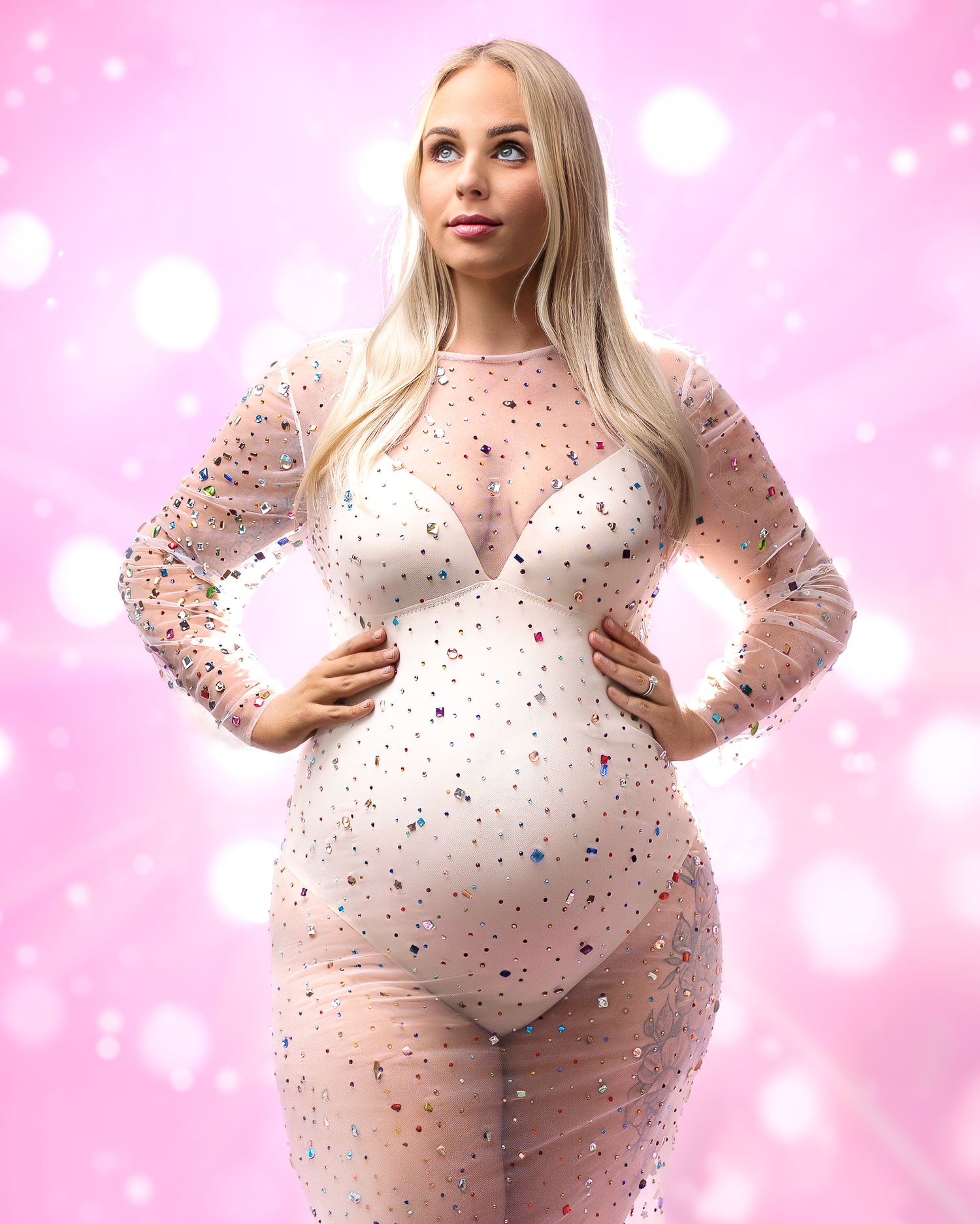 Close up shot of a pregnant lady in a sheer jewelled gown, against pink bokeh backdrop with light beams shining out behind her.