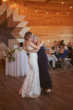 Couples slow dancing on wooden floor under warm lighting in rustic barn venue with decorative white tablecloths.