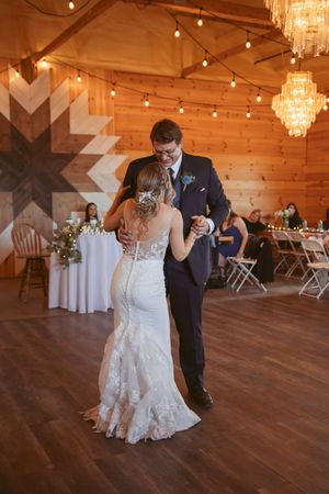 A couple dances gracefully in a rustic wooden venue decorated with elegant chandeliers and twinkling lights.