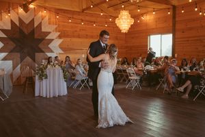 Dancers glide across a rustic wooden floor beneath warm lights in a barn venue during a romantic reception moment.