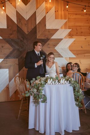 Wedding guests sit at round tables adorned with white linens and greenery in a rustic wooden barn venue.