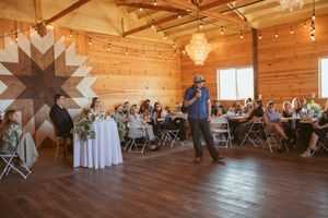 Inside a rustic wooden barn venue, guests in folding chairs watch a reception performance on a polished dance floor.