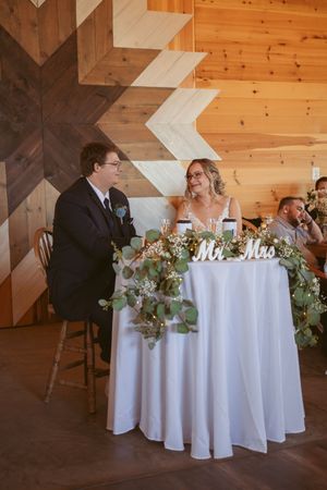 A couple sits at their sweetheart table decorated with florals in a rustic wooden venue.