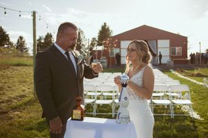 A couple shares a toast at their outdoor wedding ceremony with empty white chairs and a red barn in the background.