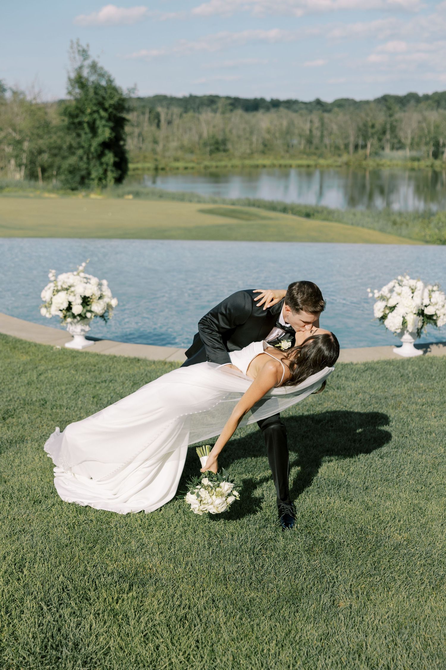 Romantic couple in wedding attire playfully dips on lush green grass near a peaceful lake during outdoor celebration.