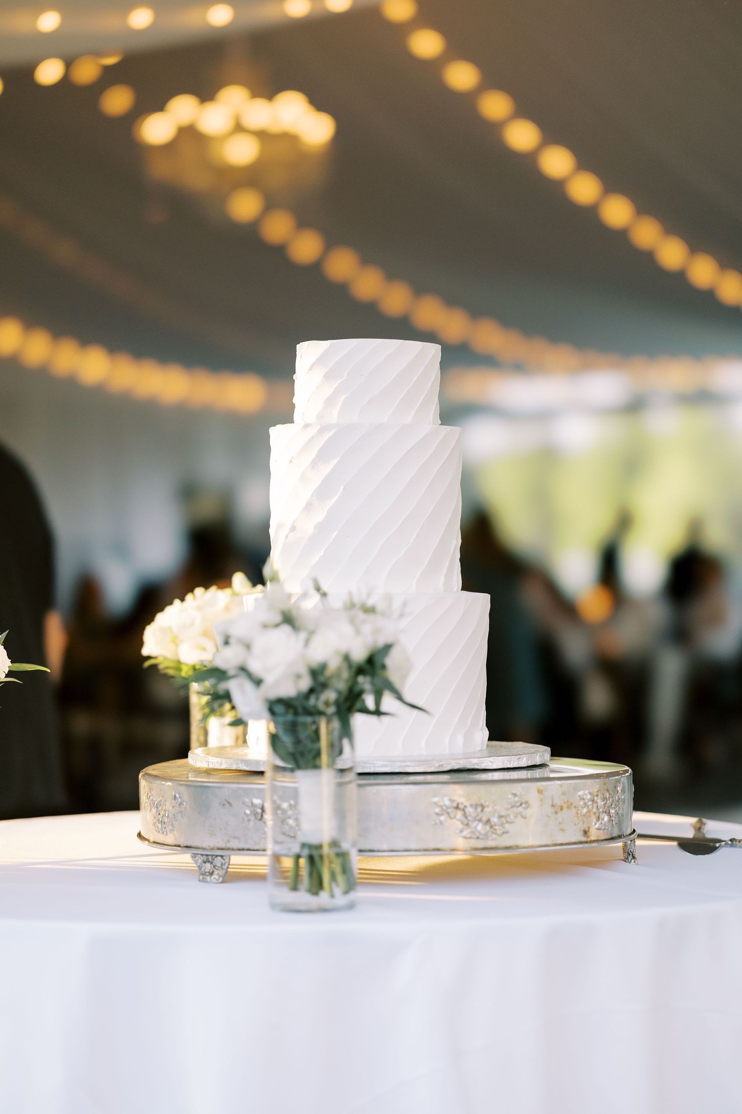 A three-tiered white wedding cake with textured frosting sits on a silver stand, decorated with small white floral arrangements.