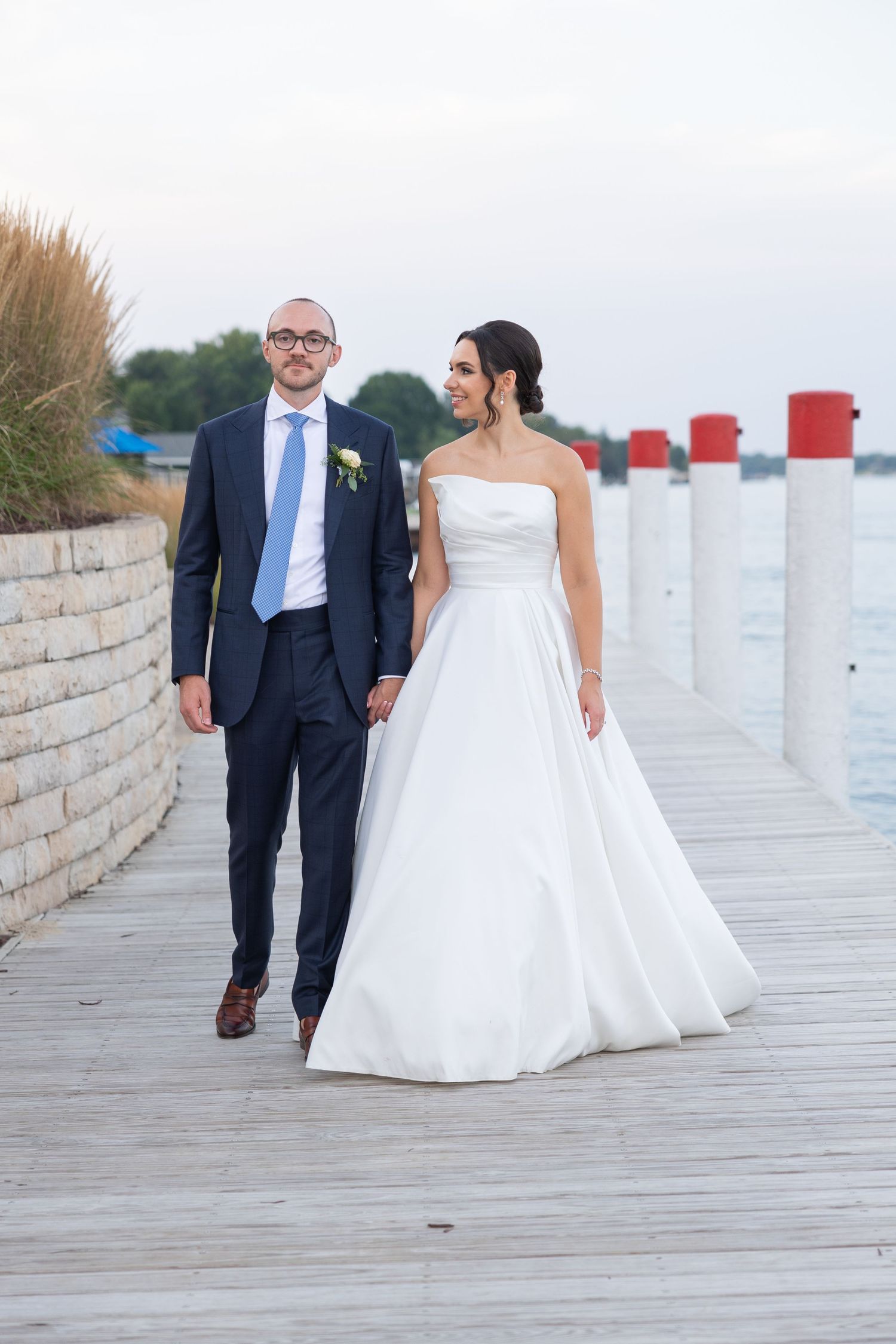 A couple in wedding attire walks together on a wooden boardwalk by the ocean at sunset during their wedding in Metro Detroit Rachel Martinez