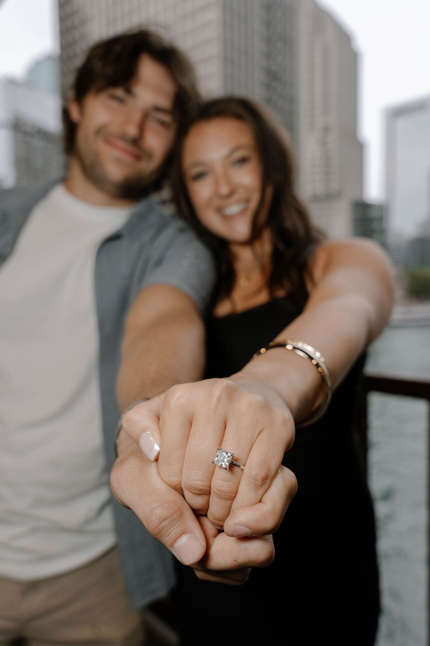 Engagement ring shown on hand with cityscape backdrop and soft-focus couple in casual attire.