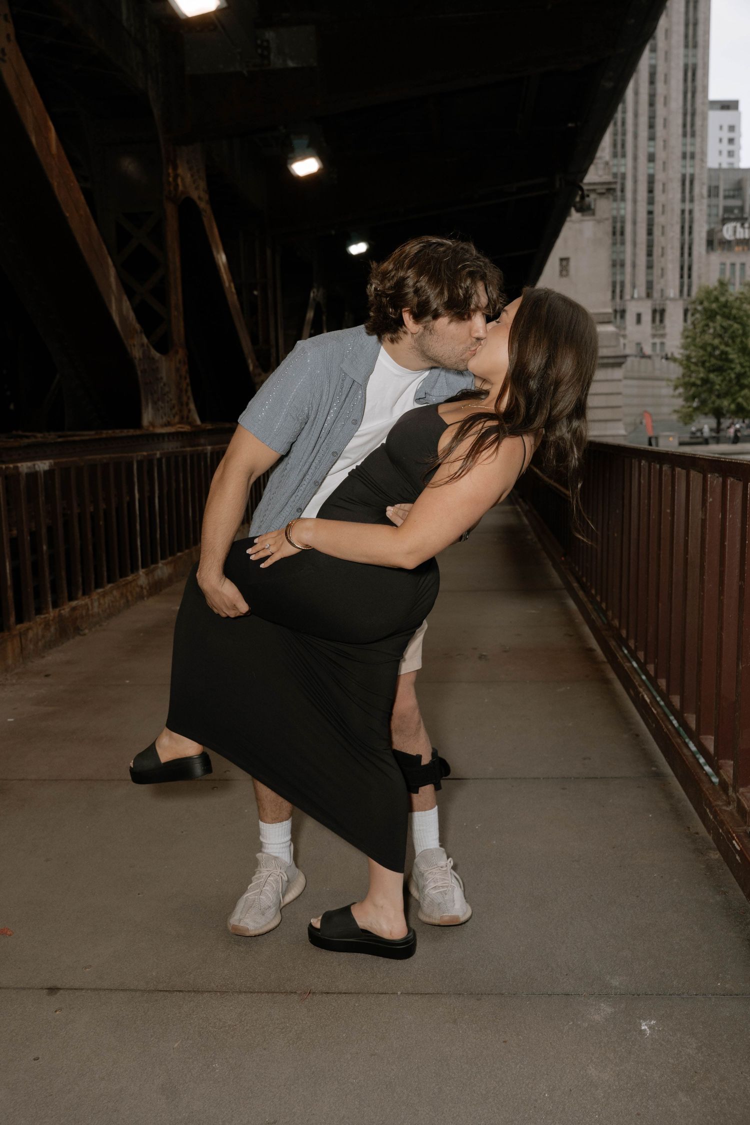 A romantic couple sharing a kiss on a covered bridge walkway at dusk with city buildings in the background.