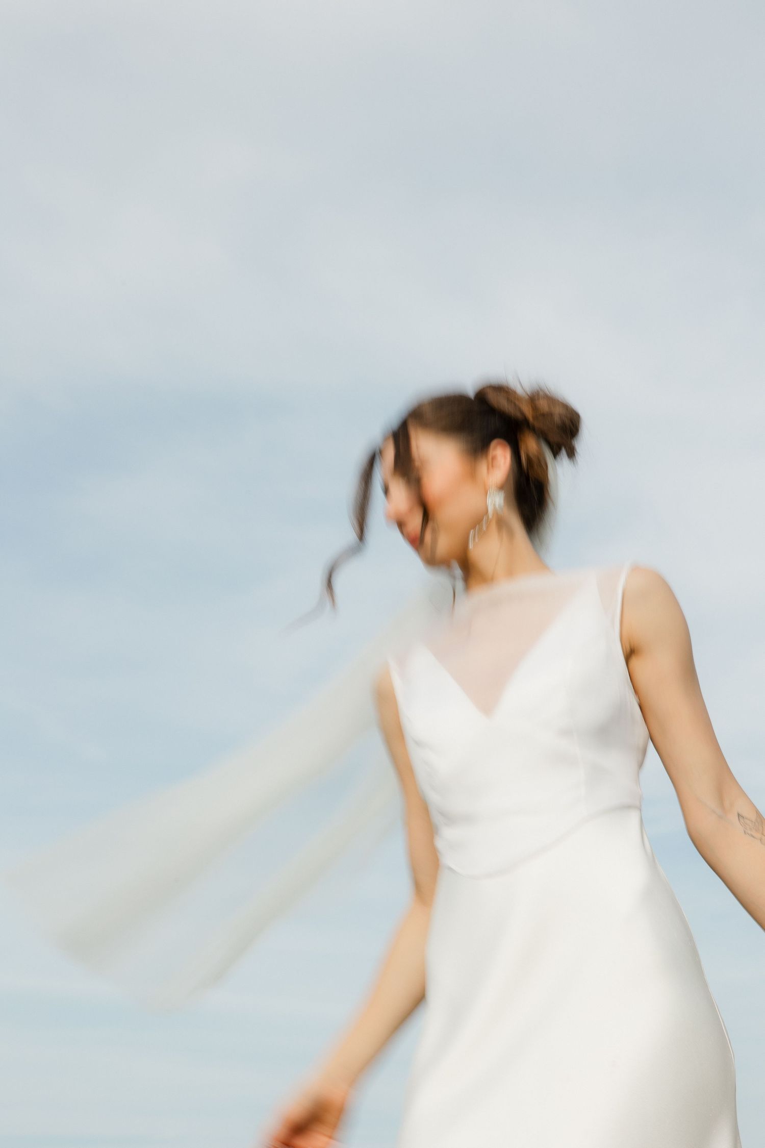 Motion-blurred bride in white gown with veil spinning against a blue sky backdrop.