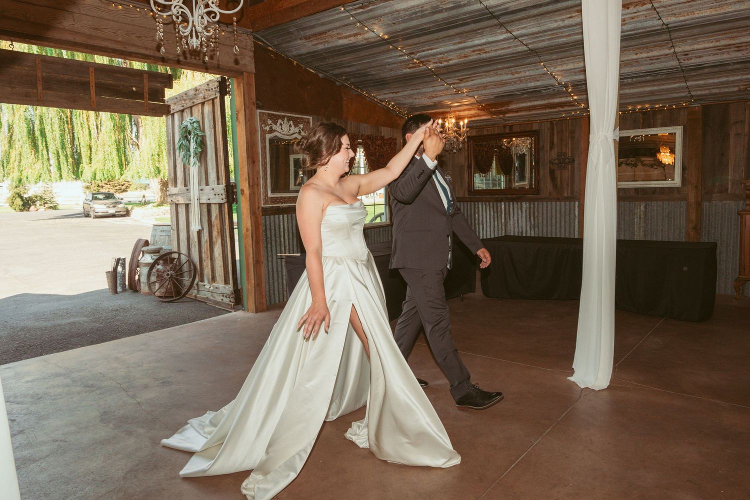 Newlyweds share their first dance in a rustic barn venue decorated with elegant chandeliers and fabric draping.