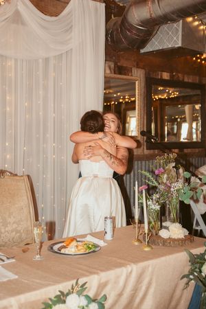 Two people share an emotional embrace at a wedding reception with fairy lights and floral centerpieces on display.