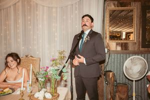 Speaker stands at rustic venue table decorated with floral centerpieces and white drapery backdrop.