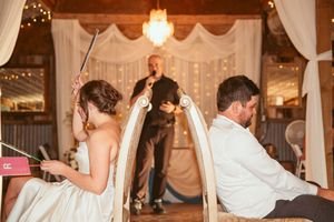 Wedding couple sits back-to-back during a reception game while an officiant stands between them.