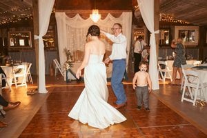 Wedding guests dance together on a rustic wooden dance floor in a barn venue decorated with white drapes and twinkling lights.