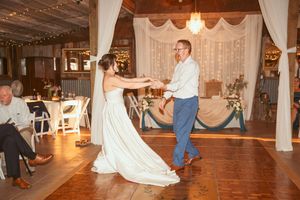 People enjoying a wedding reception with dancing on a rustic wood floor in a barn venue with hanging string lights.
