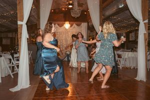 Wedding guests dance and celebrate at a rustic venue with white drapes and warm lighting during a reception party.