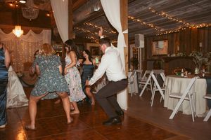 Wedding guests dance and celebrate on a wooden dance floor in a rustic barn venue decorated with string lights.