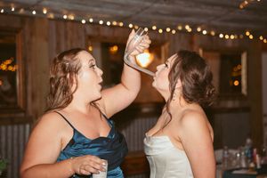 Two wedding guests share a fun moment while dancing at a rustic barn reception with string lights overhead.