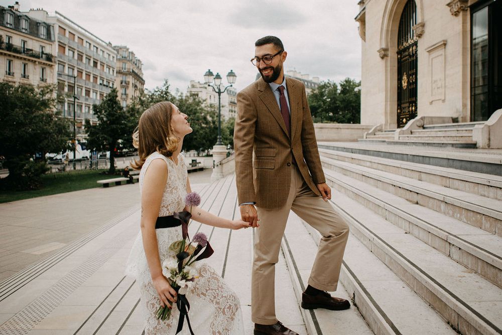 Joyful newlywed couple holding hands and smiling at each other on steps of a Parisian landmark.