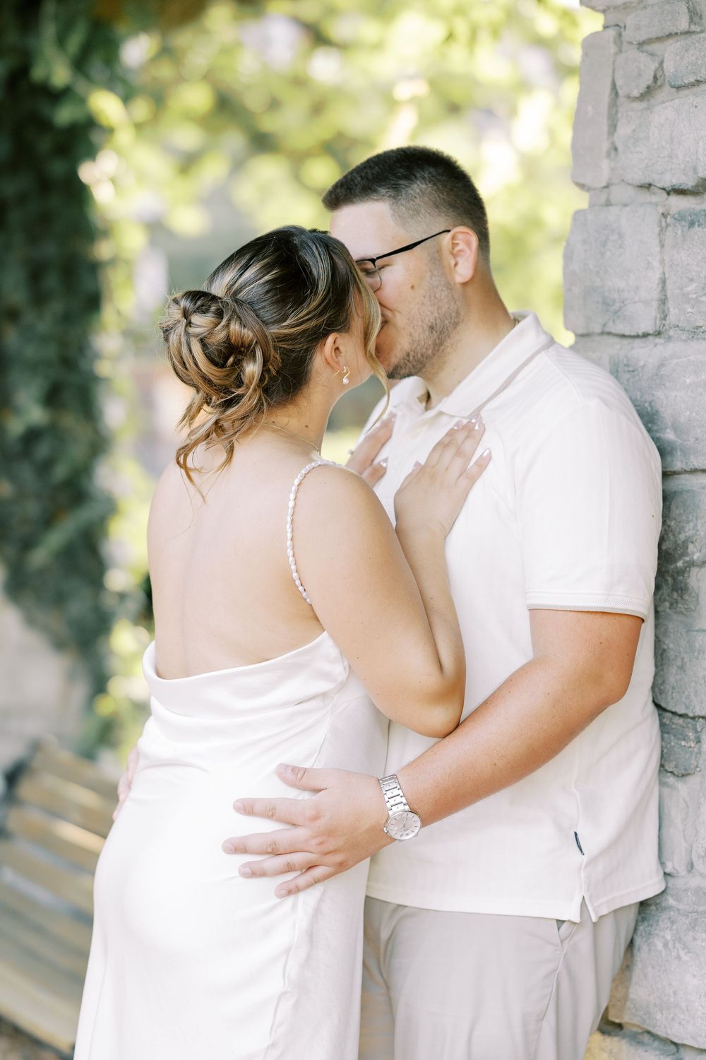 Romantic outdoor photo of a couple in white attire sharing an intimate moment against a stone wall in natural lighting.