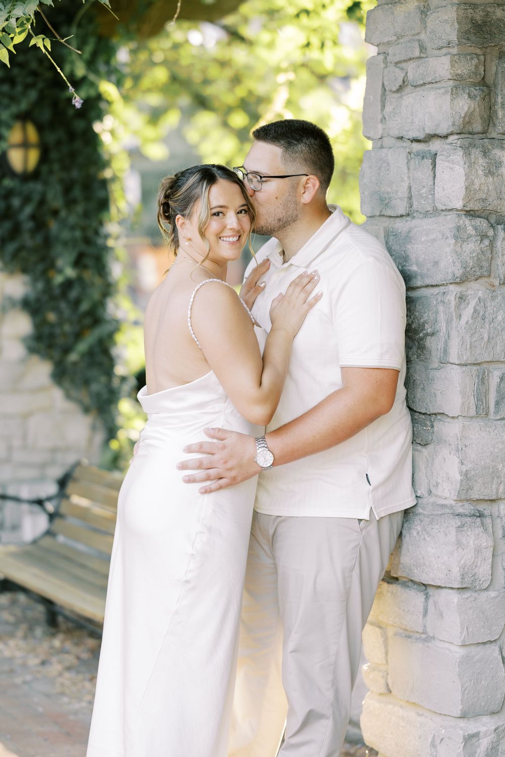 Romantic couple in white outfits sharing intimate moments against stone columns in a sunlit outdoor setting.