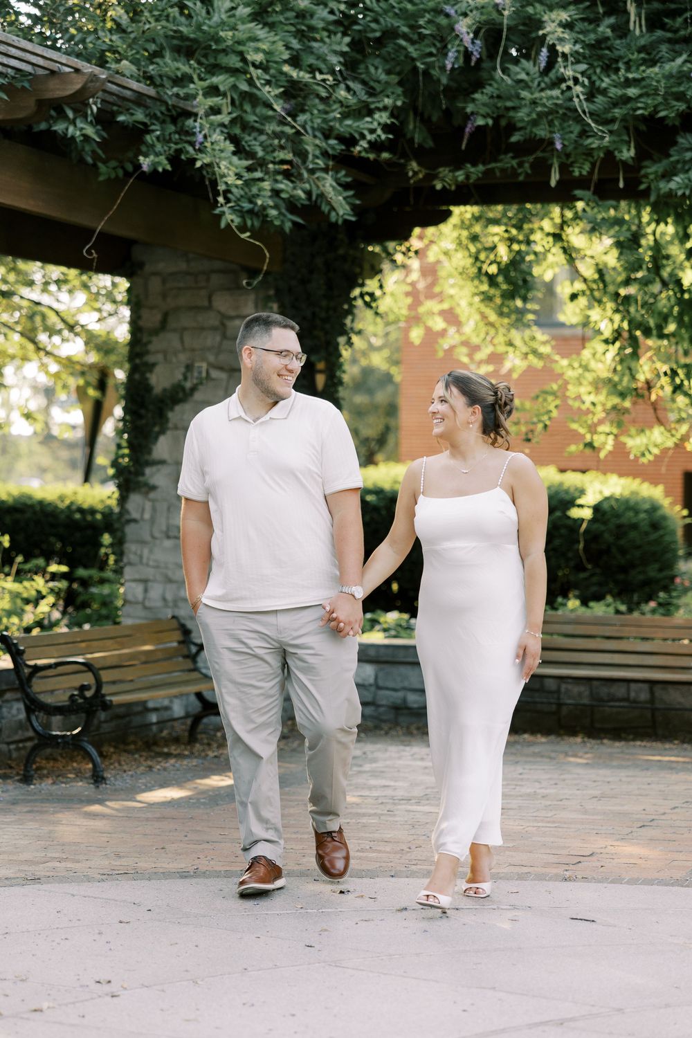 A couple dressed in white outfits walks together on a stone patio path in a garden setting during a sunny day.