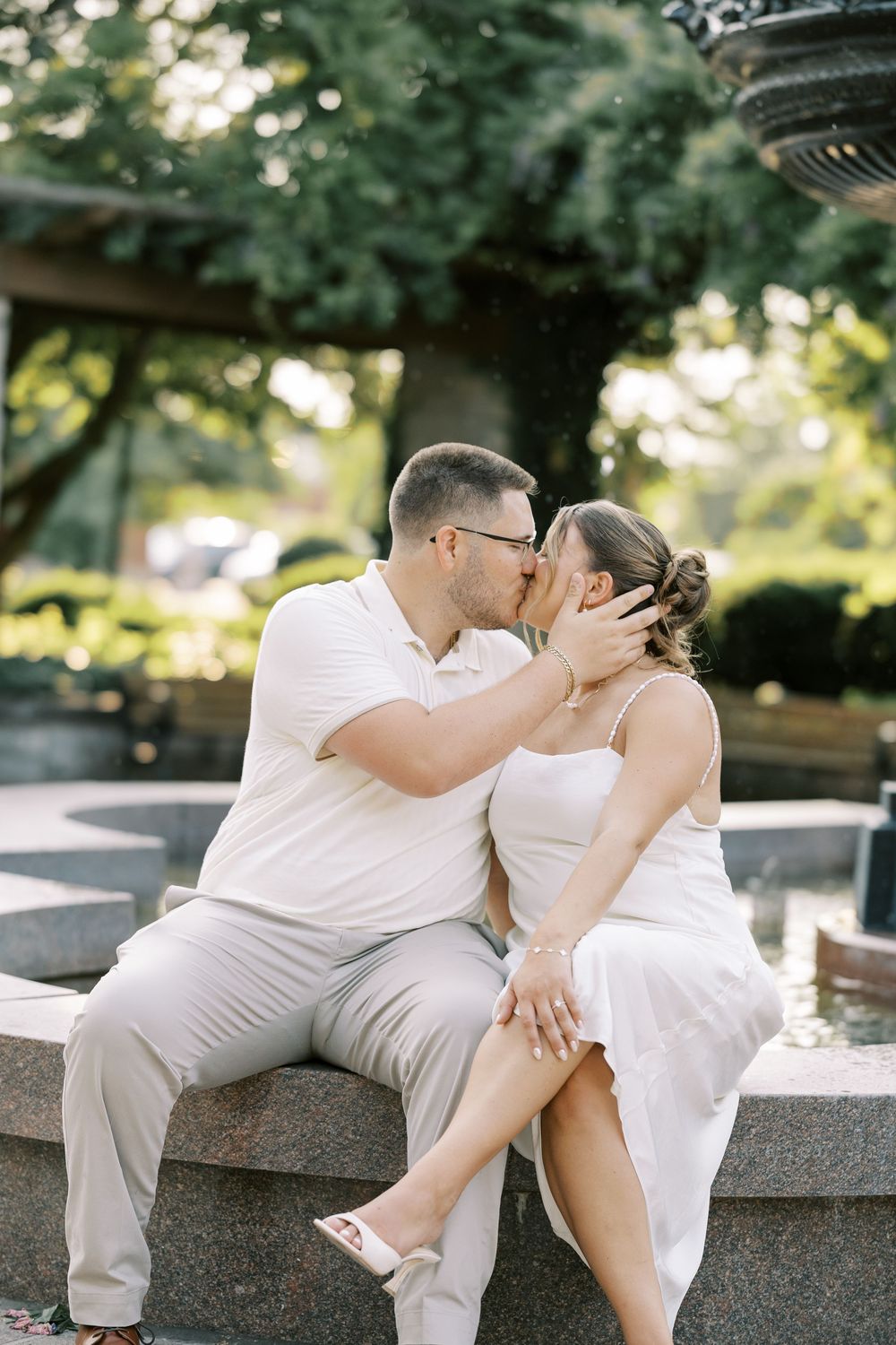 A couple in white outfits share romantic poses while sitting on a stone ledge in a park setting.