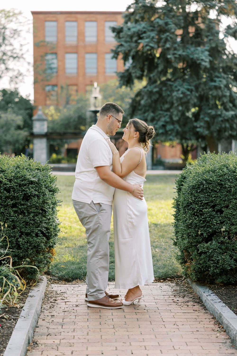 A couple in white outfits share an intimate moment on a brick pathway surrounded by manicured shrubs and trees.
