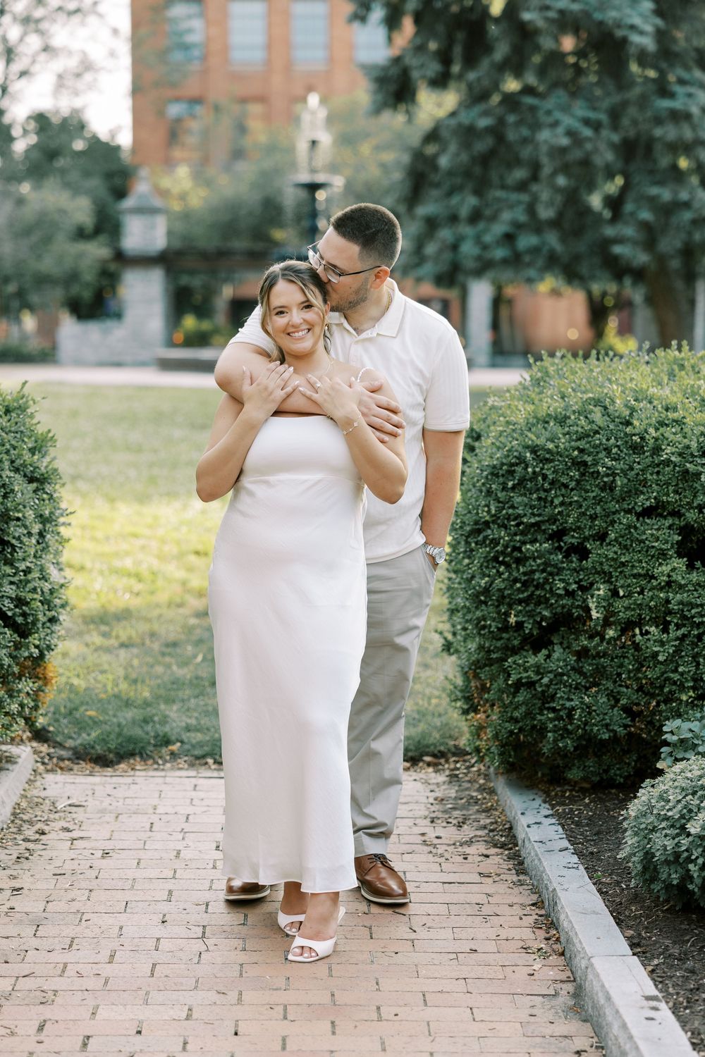 A couple in white stands on a brick pathway surrounded by manicured shrubs and greenery in a romantic scene.