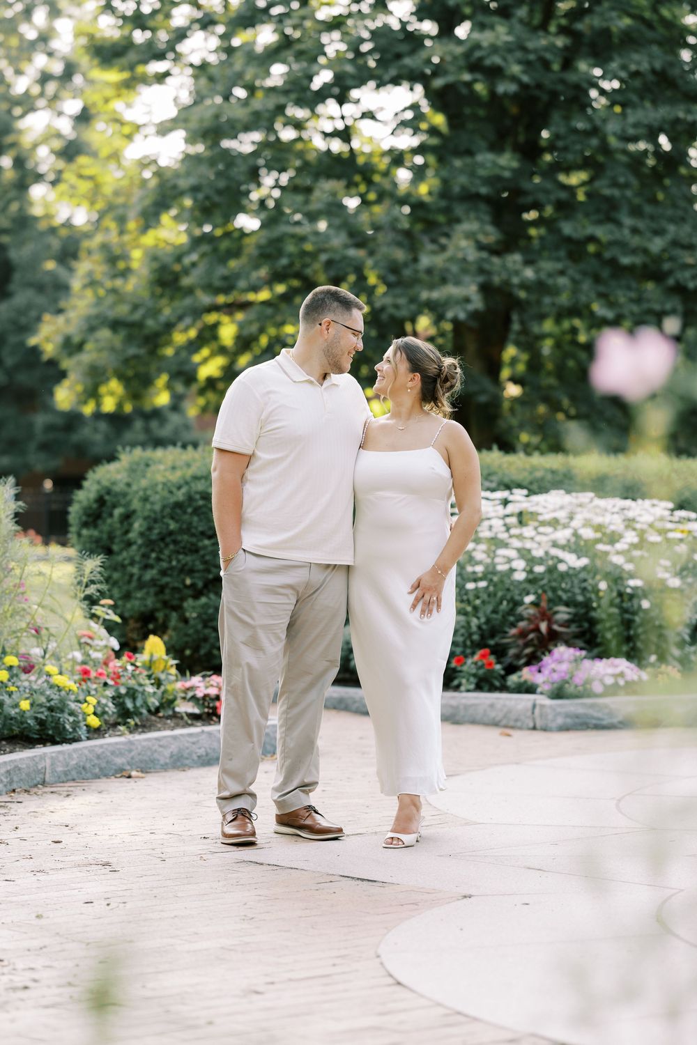 A couple in white outfits shares a romantic moment together on a garden path surrounded by blooming flowers.