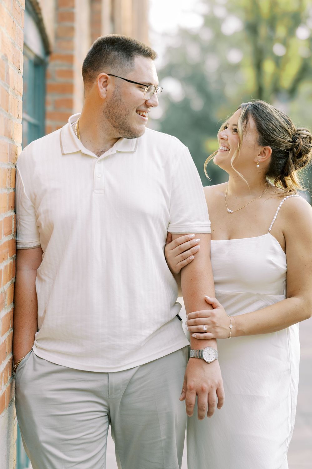 Happy couple dressed in light summer outfits share joyful moment leaning against brick wall during outdoor photoshoot.