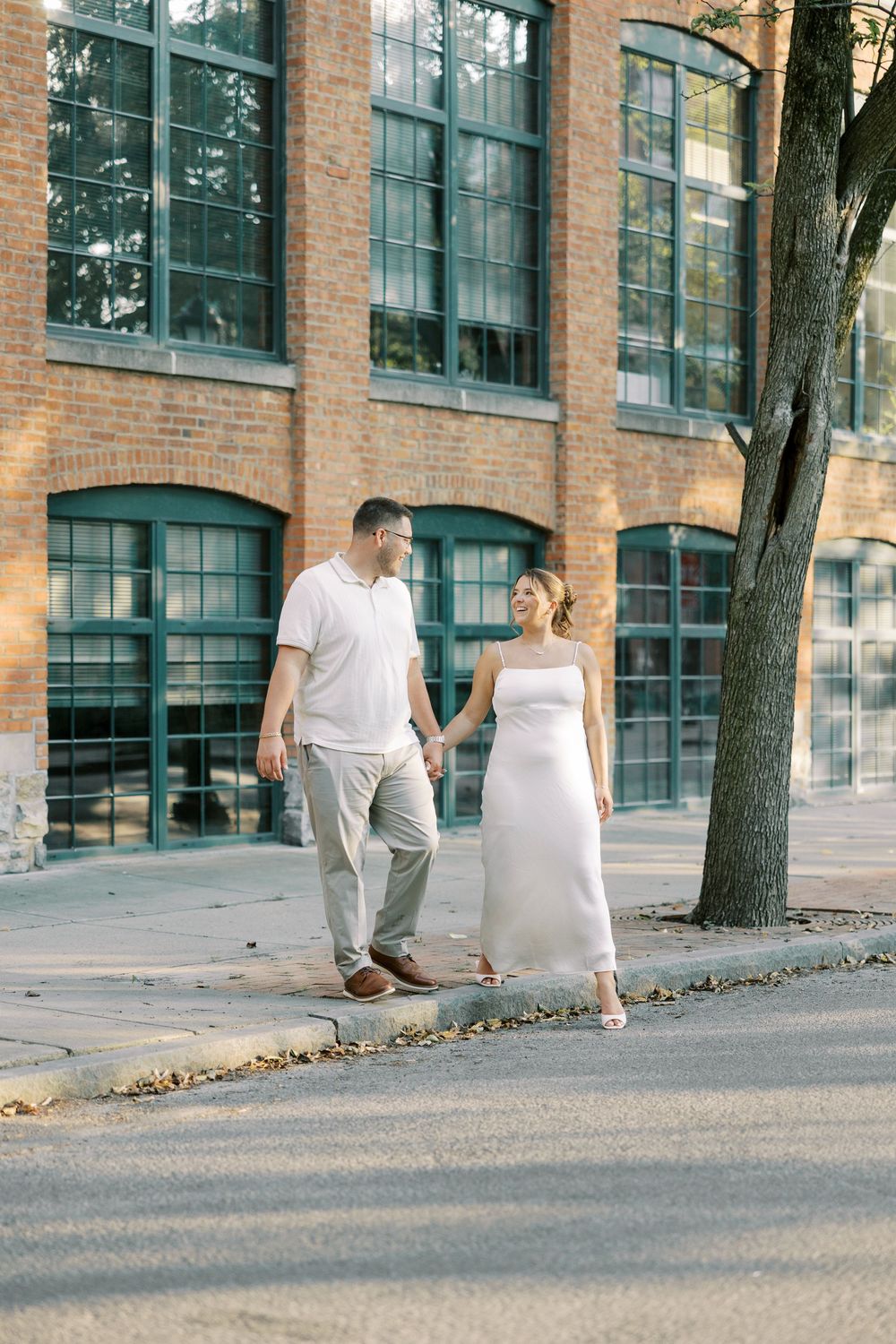 Couple in white outfits walking together down a path by a brick industrial building with large windows.