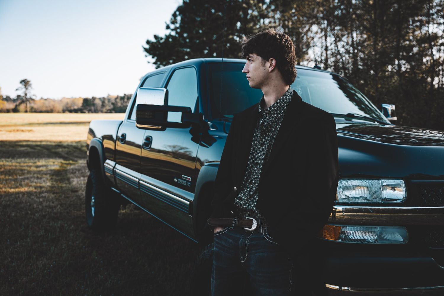 A vintage black car parked in a scenic natural setting with pine trees and open fields in the background.