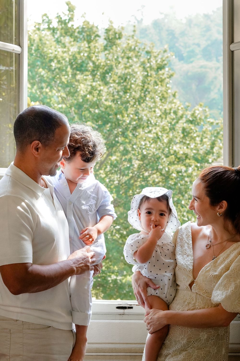 Family of four standing by open window with lush green trees, parents smiling at baby girl in white dress held by mother.